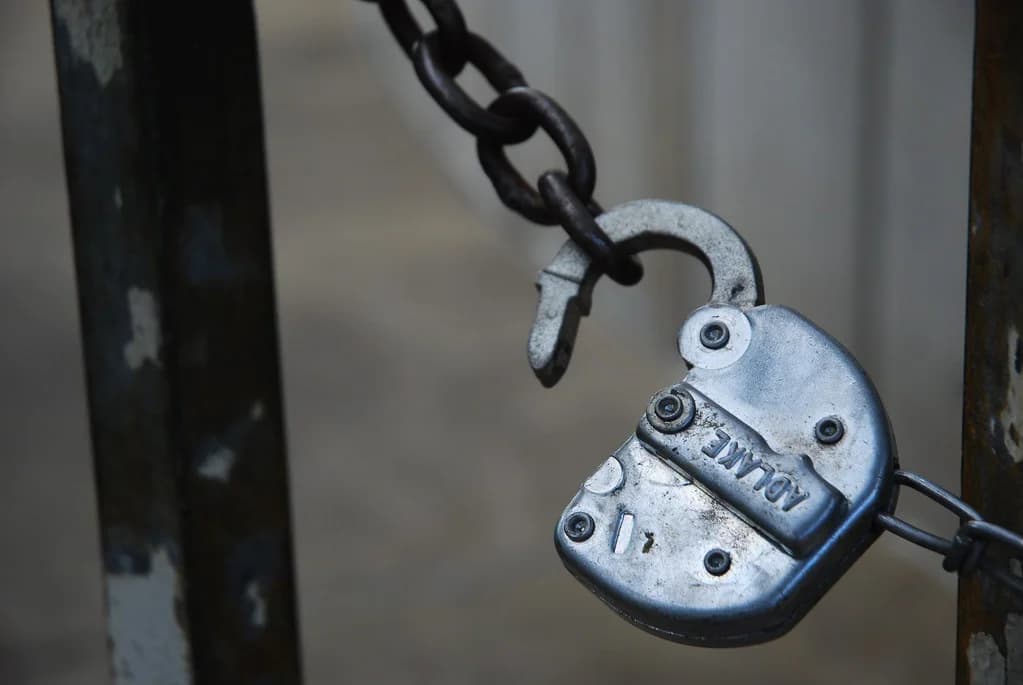 An open padlock hanging from a chain on a metal gate (photo: Tripp via Flickr, CC BY 2.0)